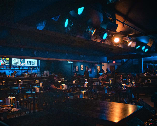 A dimly lit bar scene shows empty tables with glowing candlelit centerpieces and a bar in the background featuring a variety of drinks.