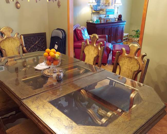 A dining table with a glass top is set with a bowl of oranges, surrounded by ornate wooden chairs in a room featuring various furnishings and decorations.