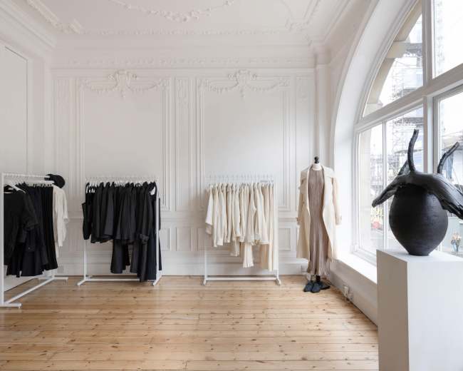 A minimalistic clothing boutique features white walls, wooden floors, and clothing racks displaying garments in neutral tones.