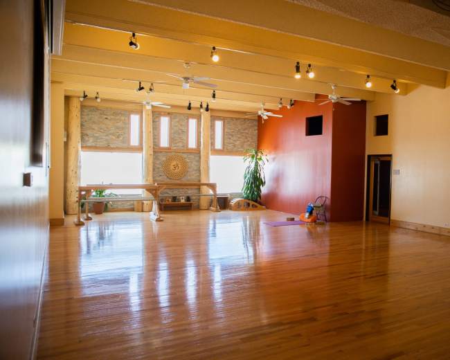 The image shows a spacious and well-lit yoga studio with wooden floors and large windows, featuring a few plants and wall decorations.
