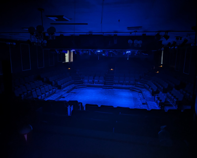 A dimly lit theater with blue lighting and empty seating surrounding a central stage area.