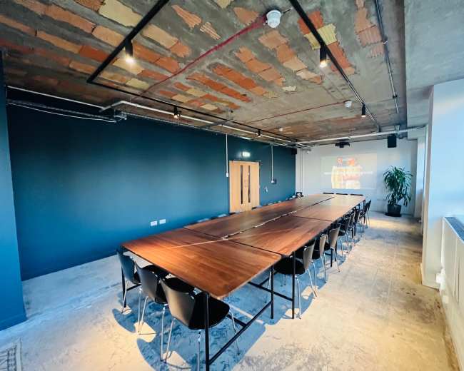 The image shows a spacious conference room featuring a long wooden table surrounded by black chairs, with a large wall projection on one side and exposed ceiling elements.