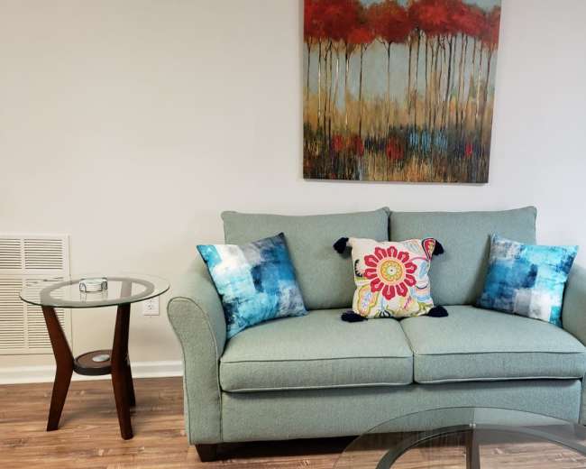The image shows a light blue couch with decorative pillows, positioned in a room featuring two glass side tables and a colorful painting of red trees on the wall.