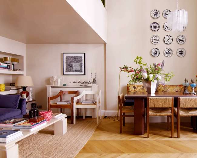 A dining area features a wooden table surrounded by chairs, adorned with a vase of flowers and decorative plates on the wall.