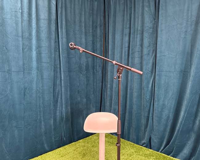 A microphone stand is positioned next to a simple chair on a grassy green surface, with a blue curtain backdrop.