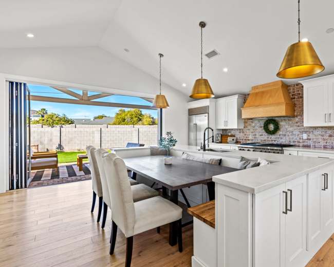 A modern kitchen features a large dining table, white cabinetry, wooden accents, and pendant lights, with a view of a backyard through sliding glass doors.