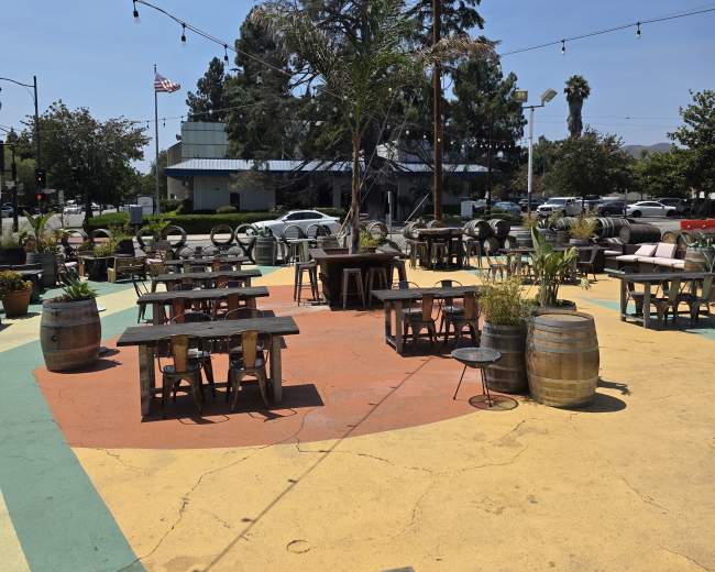 The image shows an outdoor seating area of a restaurant with tables and chairs surrounded by colorful concrete and potted plants under a clear blue sky.