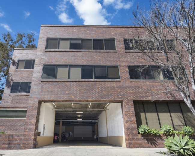 The image shows a modern brick building with large windows and an entrance to a parking area.