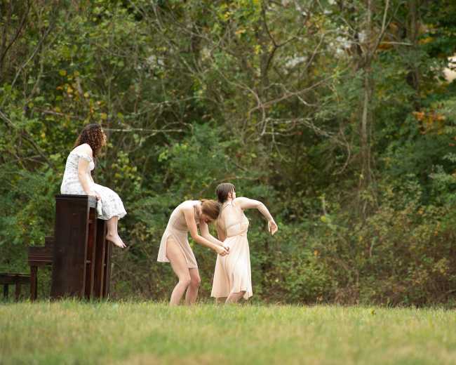 A woman sits on an upright piano while two dancers perform nearby, and a man records the scene with a camera in a natural outdoor setting.