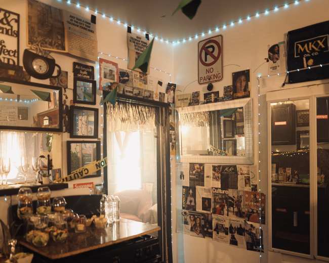 The image shows a small room with walls covered in framed posters and articles, a bar area with various items, and dim lighting accented by blue LED lights.