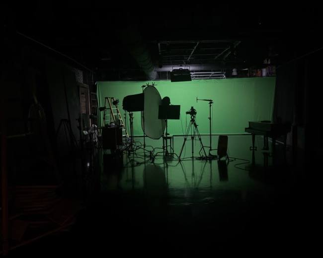 A dimly lit studio features various equipment and a large green screen as the backdrop.
