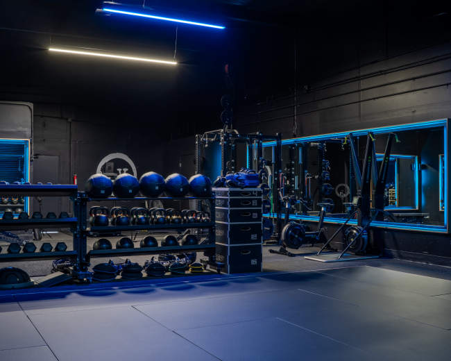 The image shows a modern gym interior with various fitness equipment, including weights and exercise mats, illuminated by blue lights.