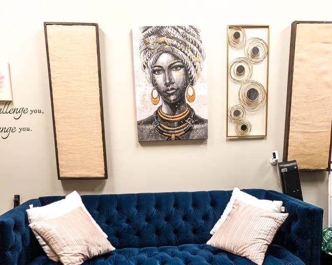 The image features a blue tufted sofa with two light-colored decorative pillows, positioned on a textured beige rug beneath a wall adorned with artistic decorations.