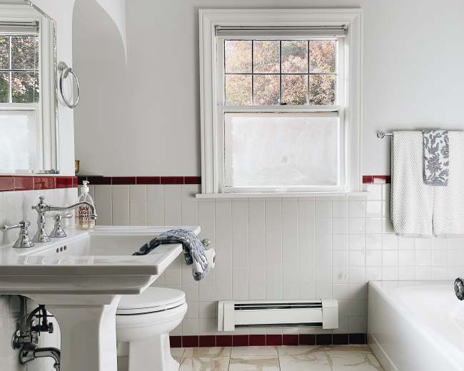 The image shows a bright bathroom with white tiled walls, a sink, a bathtub, and a window revealing a view of trees outside.