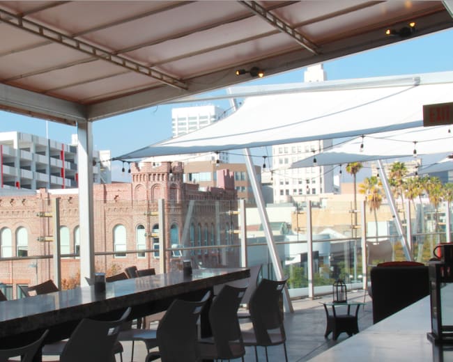 The image shows a modern rooftop bar area with a long countertop, overlooking a cityscape featuring a brick building and nearby parking structures.