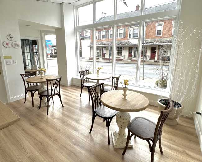 The image shows a bright, cozy café interior with wooden floors, small tables, and chairs arranged near large windows overlooking the street.