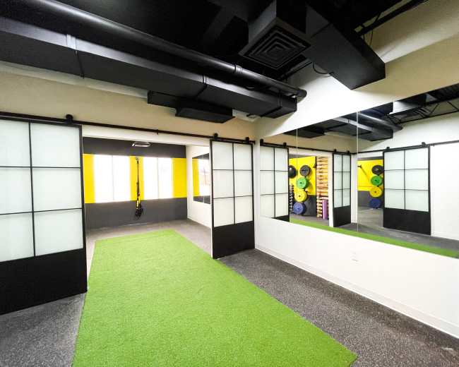 A fitness studio with a green artificial grass floor, divided by sliding black and white panel doors, and equipped with fitness equipment against a yellow wall.