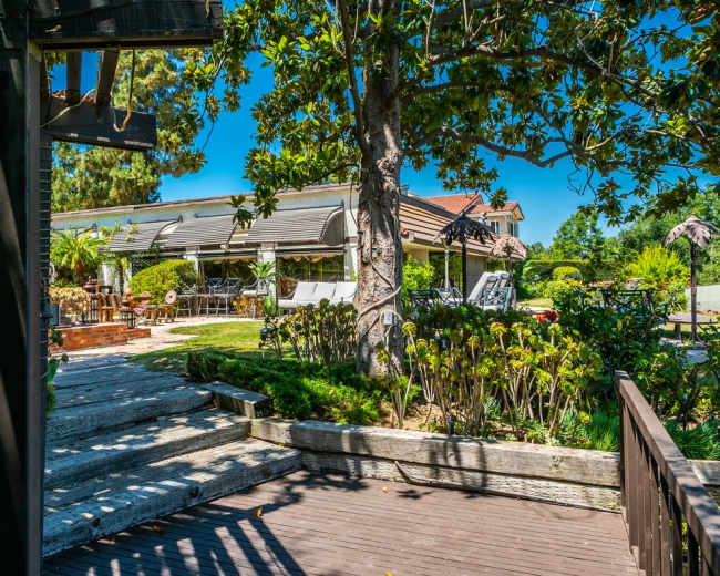 The image shows a well-maintained garden with a wooden deck, surrounded by greenery and a house with large windows in the background.