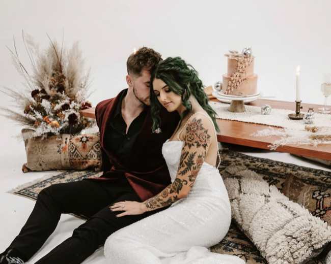 A couple sits on the floor next to a table set with a cake and decorative elements, surrounded by a rustic arrangement of plants and textiles.