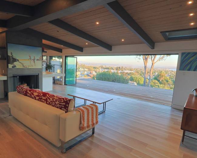 The image shows a modern, open-concept living room with a large glass door leading to an outdoor deck that offers a view of the landscape beyond.