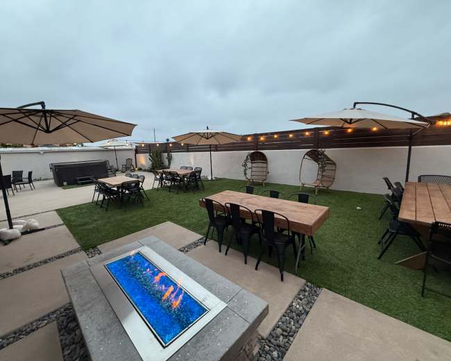 The image shows an outdoor patio area with tables, chairs, umbrellas, and a fire pit surrounded by grass and a wall.