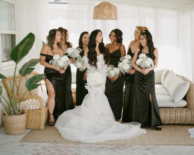 A bride stands in the center, surrounded by seven bridesmaids holding bouquets, in a bright, airy room with natural light and tropical plants.