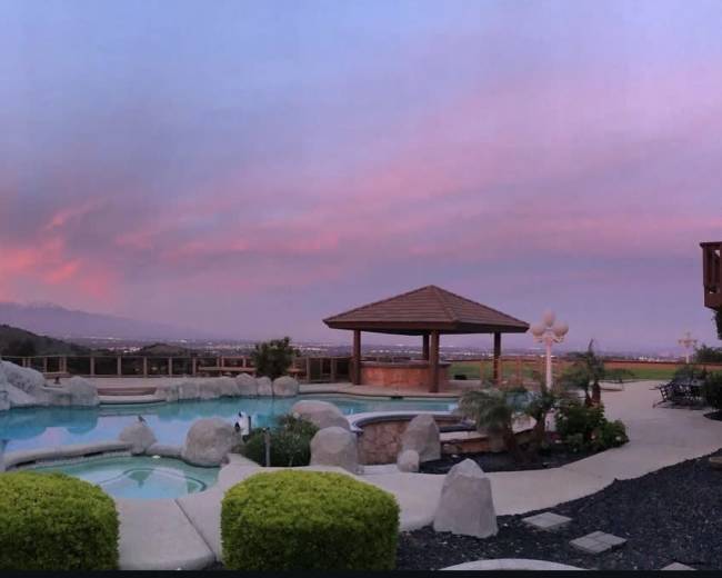 The image shows a backyard pool area with rock formations, a gazebo, and lush landscaping under a colorful sunset sky.