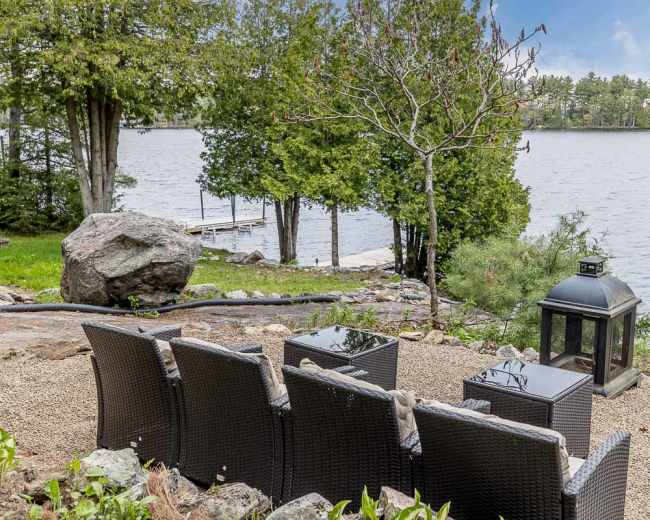 The image shows a lakeside seating area with four wicker chairs, small tables, a lantern, and a view of the water and trees.