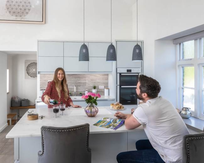 A woman serves drinks at a kitchen island while a man sits nearby, both in a modern kitchen setting.