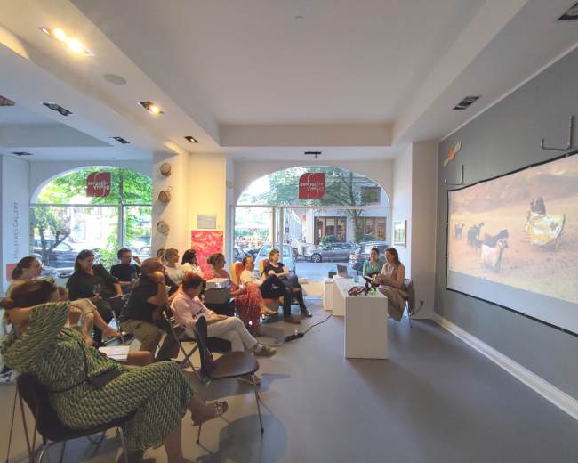 A group of people is seated in a gallery, watching a presentation that features a video projected on a wall.