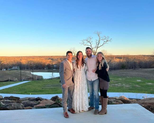 Four people stand together on a patio with a scenic landscape and a pond in the background.