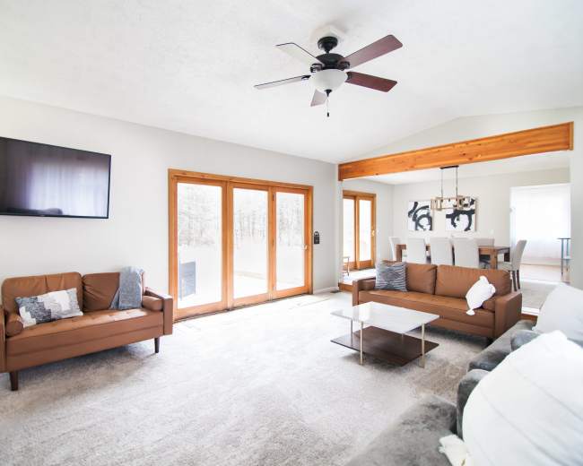 A spacious living room features two brown sofas, a glass coffee table, a wall-mounted television, and large windows that provide natural light.