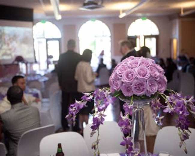 A decorative floral centerpiece featuring roses and orchids is placed on a table in a banquet hall filled with guests.