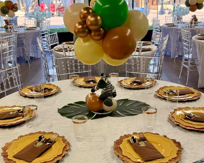 A decorated banquet room features round tables set with gold and brown tableware, surrounded by clear chairs and adorned with a colorful balloon centerpiece.
