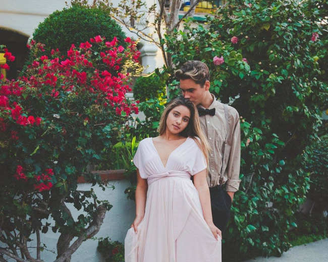 A couple stands together in front of a lush garden with vibrant flowers, the woman wearing a flowing pink dress and the man in a tan shirt.