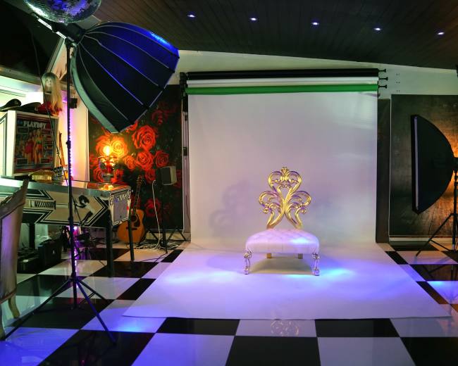 The image shows a photography studio with a white backdrop, a decorative gold chair, and various lighting equipment arranged around a black and white checkered floor.