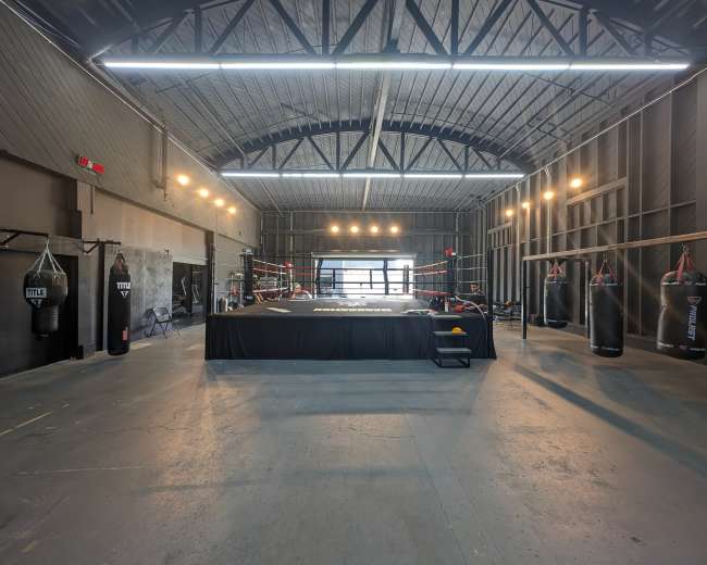 A spacious boxing gym featuring a central ring, heavy punching bags hanging from the walls, and bright overhead lighting.
