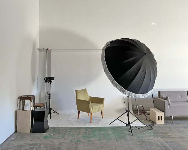 A spacious photography studio featuring a green chair, a gray sofa, a large softbox light, and various equipment against a white wall.