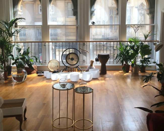 The image shows a bright room with large arched windows, featuring various plants, decorative objects, and tables on a wooden floor.