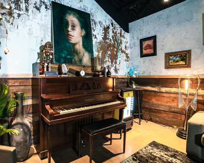 The image shows a vintage piano situated in a room with an eclectic decor, including a large painting of a girl on the wall and various artistic objects.