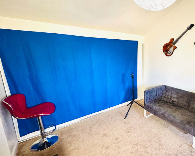 A small room featuring a blue backdrop, a gray sofa, a microphone stand, and three hanging musical instruments on the wall.