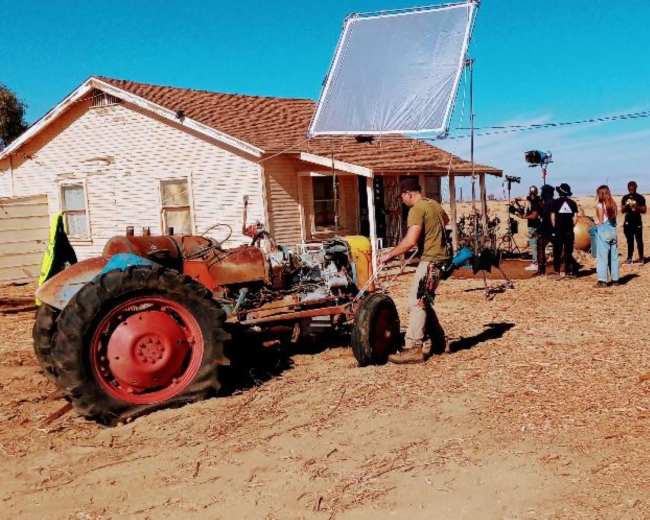 A film crew sets up equipment around a tractor and a house in a rural area.