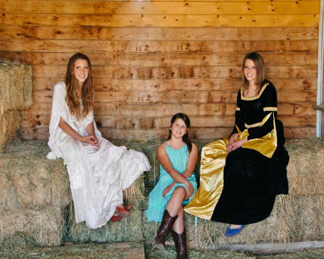 Three girls sit on hay bales inside a barn, dressed in long, flowing dresses.