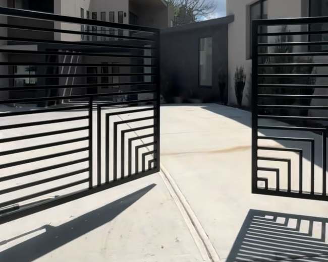 The image shows a modern entrance with open black iron gates leading to a concrete pathway and contemporary buildings in the background.