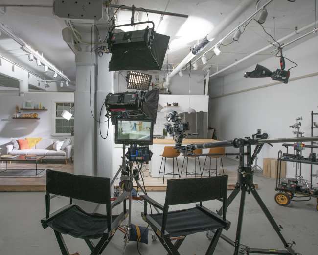 A television studio setup features two director's chairs facing a camera on a dolly, with lighting equipment above and a minimalistic living room in the background.