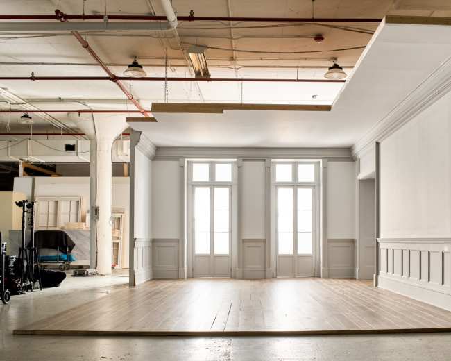 The image shows an empty studio space with wooden flooring and large windows, featuring white walls and a classic architectural design.