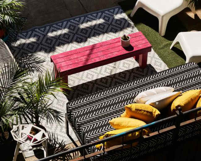 The image shows a stylish outdoor seating area with a patterned rug, a red wooden bench, a modern sofa with cushions, and white lounge chairs surrounded by plants.