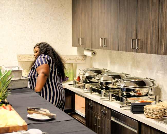 A person in a striped dress stands near a buffet table with three large chafing dishes and stacked plates in a modern kitchen.