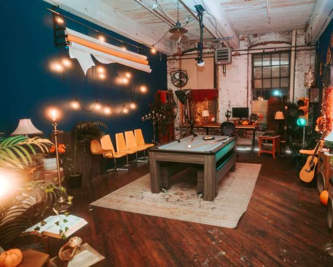 The image shows a cozy, well-decorated room featuring a pool table in the center, surrounded by various plants, furniture, and ambient lighting.