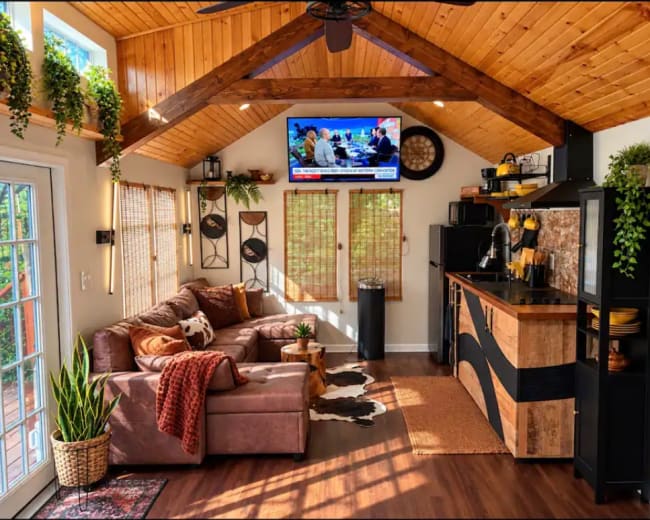 The image shows a cozy interior of a small living space with a brown sofa, a kitchen area, and a television mounted on the wall, illuminated by natural light from large windows.